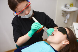 People, healthcare, modern technology, innovations and stomatology concept. Picture of woman patient in black goggles getting her teeth treated by female hygienist using dental curing light